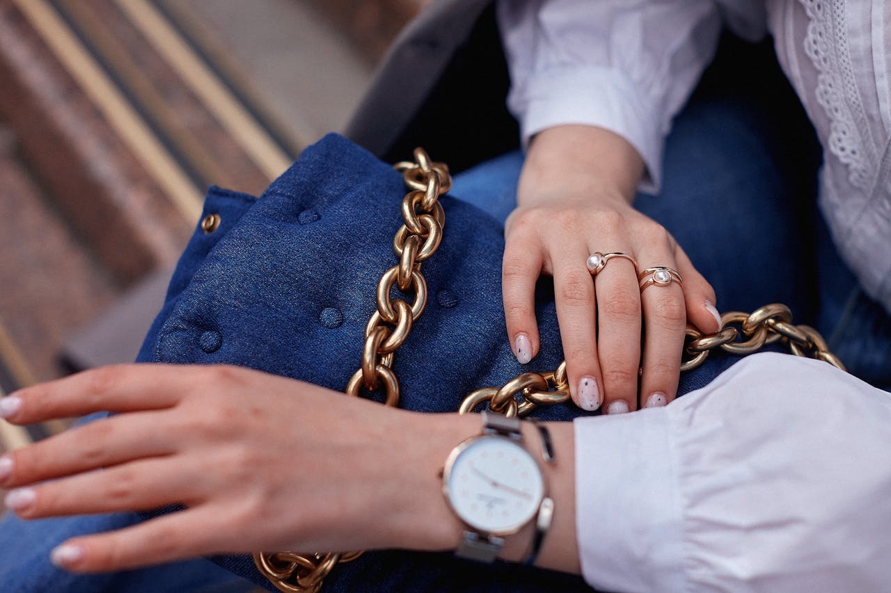 Close-up of fashionable accessories including a blue handbag with a gold chain and jewelry on hands.